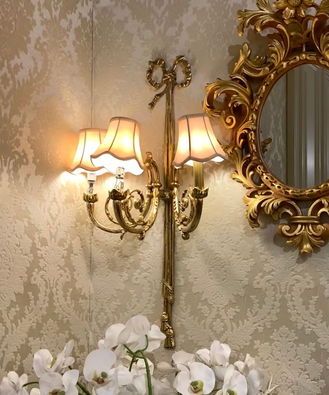 Cream damask wallpaper corner with a gilt round mirror, brass wall sconce and white orchids